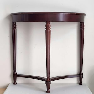 Op de afbeelding: Een donker kersenkleurige, halfronde consoletafel. De tafel heeft een gebogen blad dat wordt ondersteund door drie gefreesde poten en een lagere ligger. De poten zijn verbonden door een gebogen basis, wat een stabiel en elegant ontwerp cre&euml;ert.