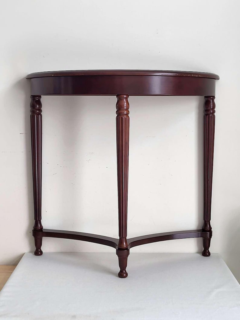 Op de afbeelding: Een donker kersenkleurige, halfronde consoletafel. De tafel heeft een gebogen blad dat wordt ondersteund door drie gefreesde poten en een lagere ligger. De poten zijn verbonden door een gebogen basis, wat een stabiel en elegant ontwerp cre&euml;ert.