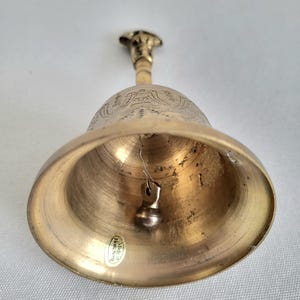 May include: A brass bell with a detailed floral design on the upper portion. The bell has a small clapper and a handle with a decorative top. The bell is made in Hong Kong.
