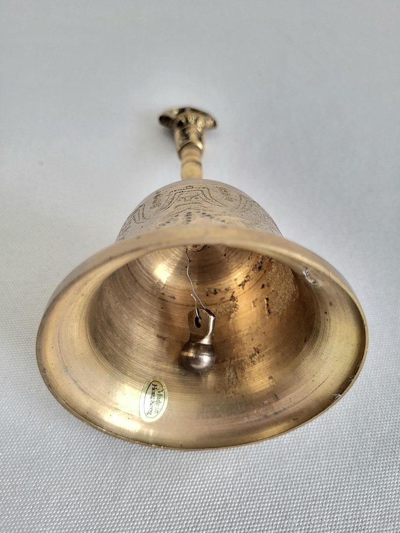 May include: A brass bell with a detailed floral design on the upper portion. The bell has a small clapper and a handle with a decorative top. The bell is made in Hong Kong.
