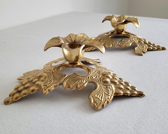 May include: Two vintage brass candle holders. Each holder features a floral cup atop a decorative base with a grape leaf design. The candle holders are made of brass and have a warm, golden color.