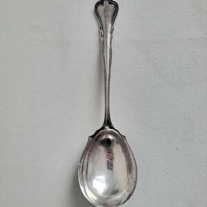 May include: A silver serving spoon with a decorative handle and a large, oval bowl. The handle features an ornate design with a monogram. The spoon is made of a shiny, reflective metal, likely silver or a silver-plated alloy. This antique spoon is suitable for serving.