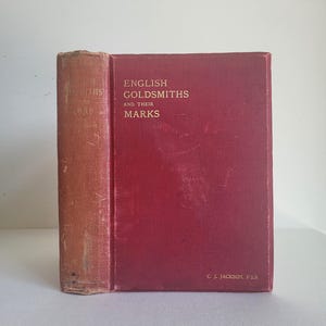 May include: A vintage book titled "English Goldsmiths and Their Marks" with a red cover and spine. The spine is a faded tan colour. The book's title is in gold lettering. The author's name, C.J. Jackson, F.S.A., is printed at the bottom.
