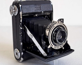 Peut inclure: Un appareil photo vintage noir Zeiss Ikon Derval avec un objectif et des accents argentés. L'appareil photo est ouvert, révélant l'objectif et le soufflet. L'objectif porte l'inscription "Zeiss Ikon" et "Nettar-Anastigmat". L'appareil photo est sur une surface blanche.