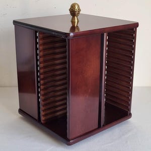 May include: A square, dark brown wooden media storage rack with a rotating base. The unit features four sides with horizontal slots for organizing discs or cassettes. A small, decorative brass-colored finial sits centered on the flat top surface.