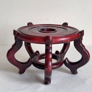 Peut inclure: Un présentoir en bois rouge foncé avec un dessus circulaire et des pieds courbes ornés. Le présentoir a une ouverture centrale et un anneau inférieur, probablement pour tenir un objet décoratif. Le présentoir mesure environ 15 cm de diamètre.
