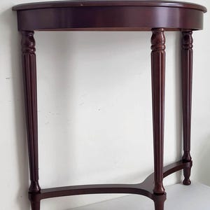 Op de afbeelding: Een donker kersenkleurig gebeitste, halvemaanvormige consoletafel. De tafel heeft een afgerond blad dat wordt ondersteund door vier gedraaide poten met decoratieve details. De poten zijn verbonden door een gebogen ligger. De tafel is ontworpen om tegen een muur te worden geplaatst.