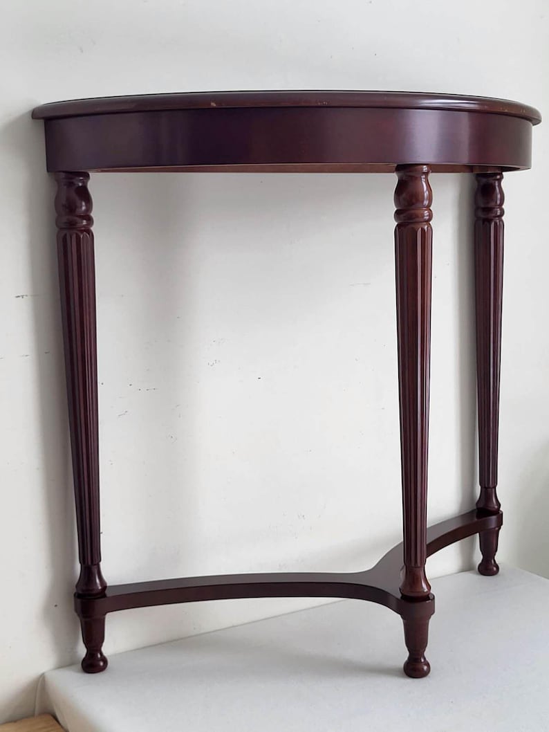 Op de afbeelding: Een donker kersenkleurig gebeitste, halvemaanvormige consoletafel. De tafel heeft een afgerond blad dat wordt ondersteund door vier gedraaide poten met decoratieve details. De poten zijn verbonden door een gebogen ligger. De tafel is ontworpen om tegen een muur te worden geplaatst.