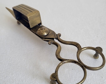 May include: Antique brass scissors with a rectangular blade guard. The scissors have ornate handles with finger holes and a small, pointed blade. The metal has a warm, aged patina, suggesting vintage craftsmanship. The scissors are likely for fabric or paper.