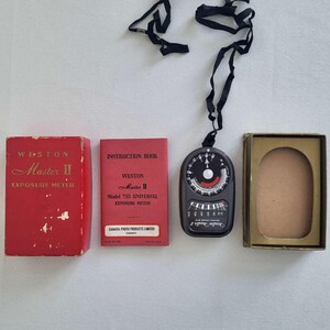 Peut inclure: Un kit de posemètre Weston Master II vintage. Le kit comprend une boîte rouge, un livret d'instructions rouge, un posemètre noir avec une sangle noire et une boîte dorée. Le posemètre a un cadran et divers réglages.