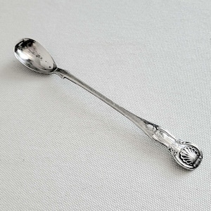 May include: A long-handled silver-toned spoon featuring an oval bowl and a decorative handle end with a raised shell motif and intricate scrollwork detailing. The metal surface exhibits a polished finish with subtle reflections along the stem.