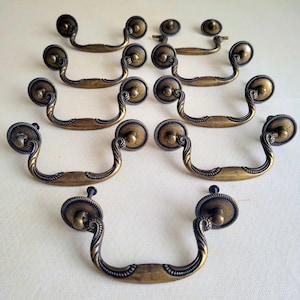 Peut inclure: Ensemble de poignées de tiroir en laiton antique. Chaque poignée présente une plaque arrière décorative et une poignée incurvée avec des détails complexes. La quincaillerie a une esthétique vintage, idéale pour la restauration de meubles.