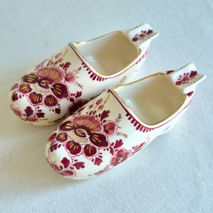 May include: Two cream-colored ceramic clogs featuring a decorative floral pattern in deep red and gold. Each shoe-shaped vessel has a smooth, glossy finish with gold-trimmed edges and a detailed botanical motif painted on the front and sides.