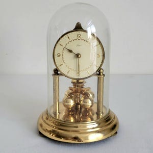 May include: A vintage gold-toned clock with a cream-colored face, encased in a clear glass dome. The clock features a pendulum with multiple gold spheres and is set on a round base. The clock's face has black numerals and hands.