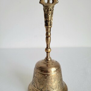 May include: A vintage brass bell with an ornate handle and a detailed, engraved design on the bell's body. The handle features a decorative top and a carved face. The bell is a warm, golden color.