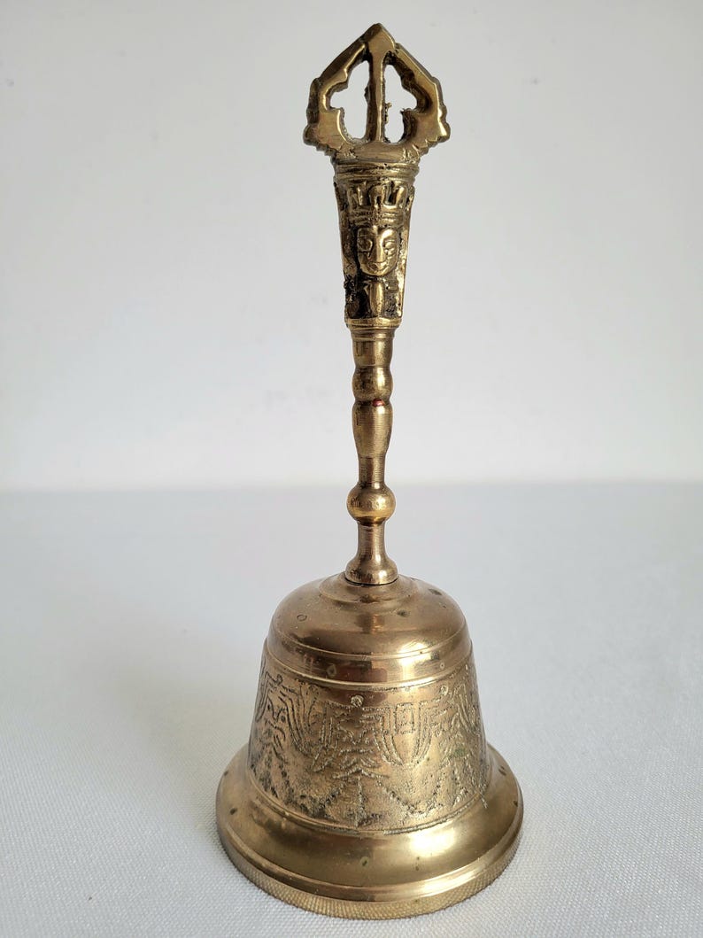 May include: A vintage brass bell with an ornate handle and a detailed, engraved design on the bell's body. The handle features a decorative top and a carved face. The bell is a warm, golden color.