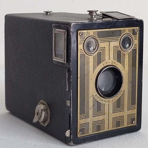 May include: A vintage black Kodak Brownie Junior Six-20 box camera. The camera features a decorative gold-coloured faceplate with geometric patterns and the text "SIX-20" and "BROWNIE JUNIOR". It has a viewfinder and a winding key.