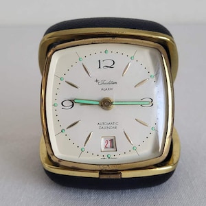 May include: A vintage Tradition alarm clock with a cream-colored face, gold-tone frame, and black leatherette case. The clock face features black numerals, green accents, and a calendar displaying the number 27. The words "Tradition" and "Alarm" are printed on the face.