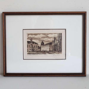 May include: A framed black and white etching depicting a cityscape with historical buildings, including a church with a tall spire. The print is titled Budapest Hilton 1980, numbered 144/200, and signed by the artist. It features a dark wood frame.