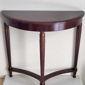 Op de afbeelding: Een halfronde console in donker kersenhout. De tafel heeft een halfrond blad dat wordt ondersteund door drie gefreesde poten, verbonden door een gebogen ligger. De tafel is ongeveer 76 cm breed en 38 cm diep.