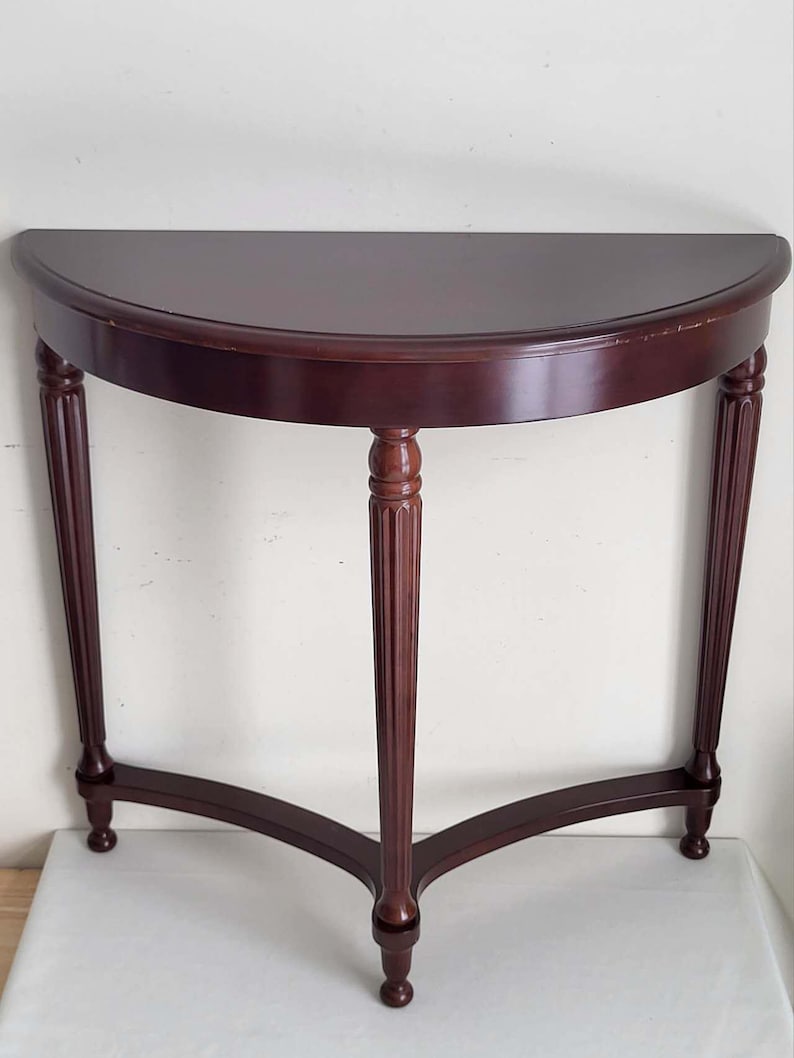 Op de afbeelding: Een halfronde console in donker kersenhout. De tafel heeft een halfrond blad dat wordt ondersteund door drie gefreesde poten, verbonden door een gebogen ligger. De tafel is ongeveer 76 cm breed en 38 cm diep.