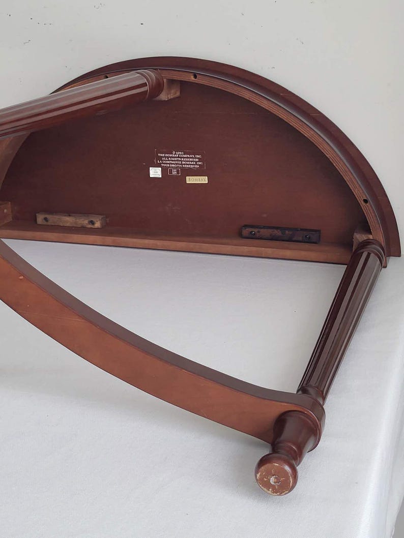 Op de afbeelding: Een halfronde houten tafel met twee gebogen en twee rechte poten. De tafel is donkerbruin van kleur met een glanzende afwerking. De poten zijn een donkerdere bruintint. De onderkant van de tafel heeft een label.