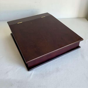 Peut inclure: Une boîte rectangulaire en bois brun foncé avec un couvercle plat et lisse et deux petites charnières en métal doré visibles sur le bord arrière. La boîte présente une base à gradins avec une section inférieure légèrement plus large.