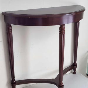 Op de afbeelding: Een halfronde console van donker kersenhout. De tafel heeft een plat blad en wordt ondersteund door drie gecanneleerde poten. De poten zijn verbonden door een gebogen ligger. De tafel is ongeveer 76 cm breed.