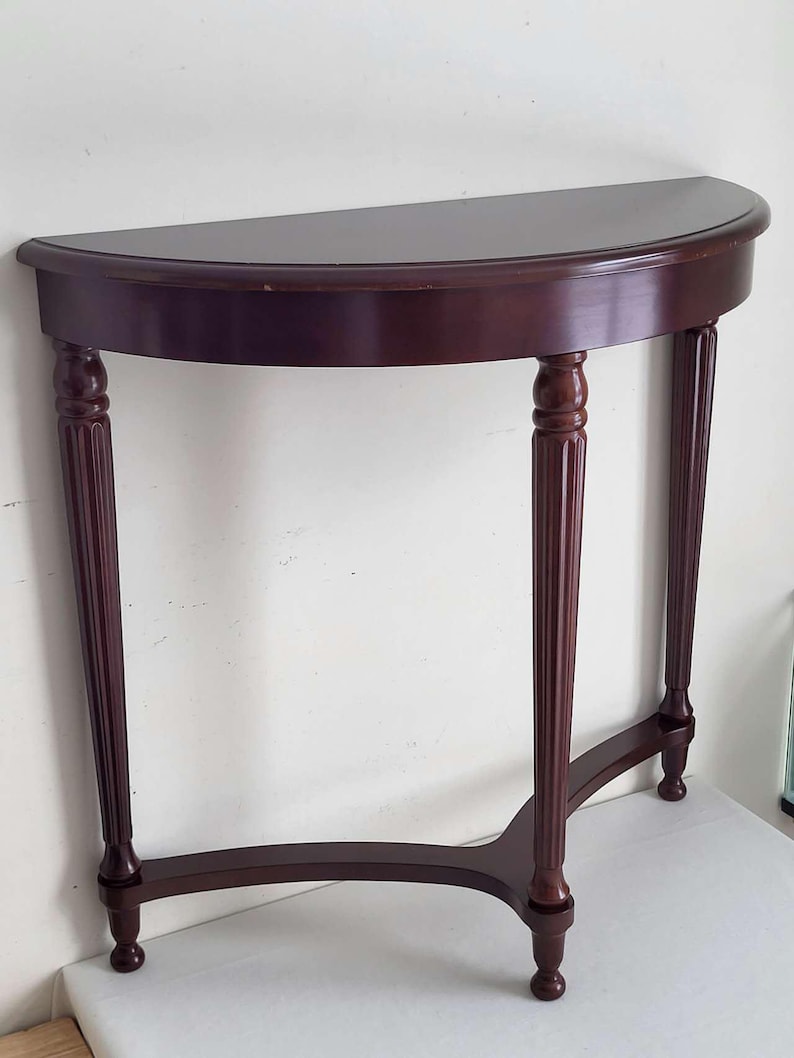 Op de afbeelding: Een halfronde console van donker kersenhout. De tafel heeft een plat blad en wordt ondersteund door drie gecanneleerde poten. De poten zijn verbonden door een gebogen ligger. De tafel is ongeveer 76 cm breed.