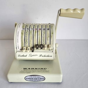 May include: An off-white vintage Paymaster cash register with a crank handle. The register displays dollar and cent amounts, with the text "Locked Paymaster Protection" and "Warning" visible. The register has a cream-colored handle.