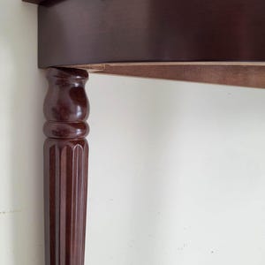 Op de afbeelding: Een gedeelte van een donkerbruine houten tafel met een gebogen rand. De tafelpoten hebben een cannelure kolomontwerp. De tafel staat tegen een witte muur.