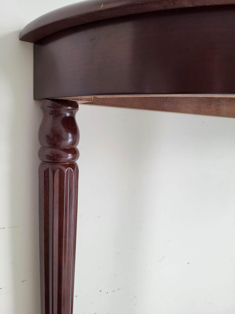 Op de afbeelding: Een gedeelte van een donkerbruine houten tafel met een gebogen rand. De tafelpoten hebben een cannelure kolomontwerp. De tafel staat tegen een witte muur.