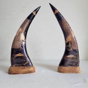 May include: Two curved, dark-coloured animal horns mounted on light-coloured wooden bases. Each horn features intricate, light-toned carvings of a bird of prey with detailed feathers and talons, along with small, decorative geometric patterns near the base.