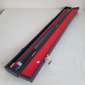 May include: A black pool cue in a black case with a red felt interior. The cue has a black and grey textured grip and a white tip. The case is open, revealing the cue and a separate red shaft. The case is rectangular with metal clasps.