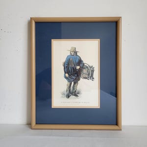 May include: Framed print of a person in a blue coat and straw hat, carrying a basket. The print is set against a navy blue mat and framed in a gold-toned wood frame. The text at the bottom reads "LE MARCHANT D'OISEAUX DE NEVADA."