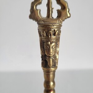 May include: A detailed, antique brass vajra, a ritual object in Buddhism and Hinduism. The vajra features intricate carvings, including a face and ornate crown. The handle has a series of rounded sections, and the top has a three-pronged design.