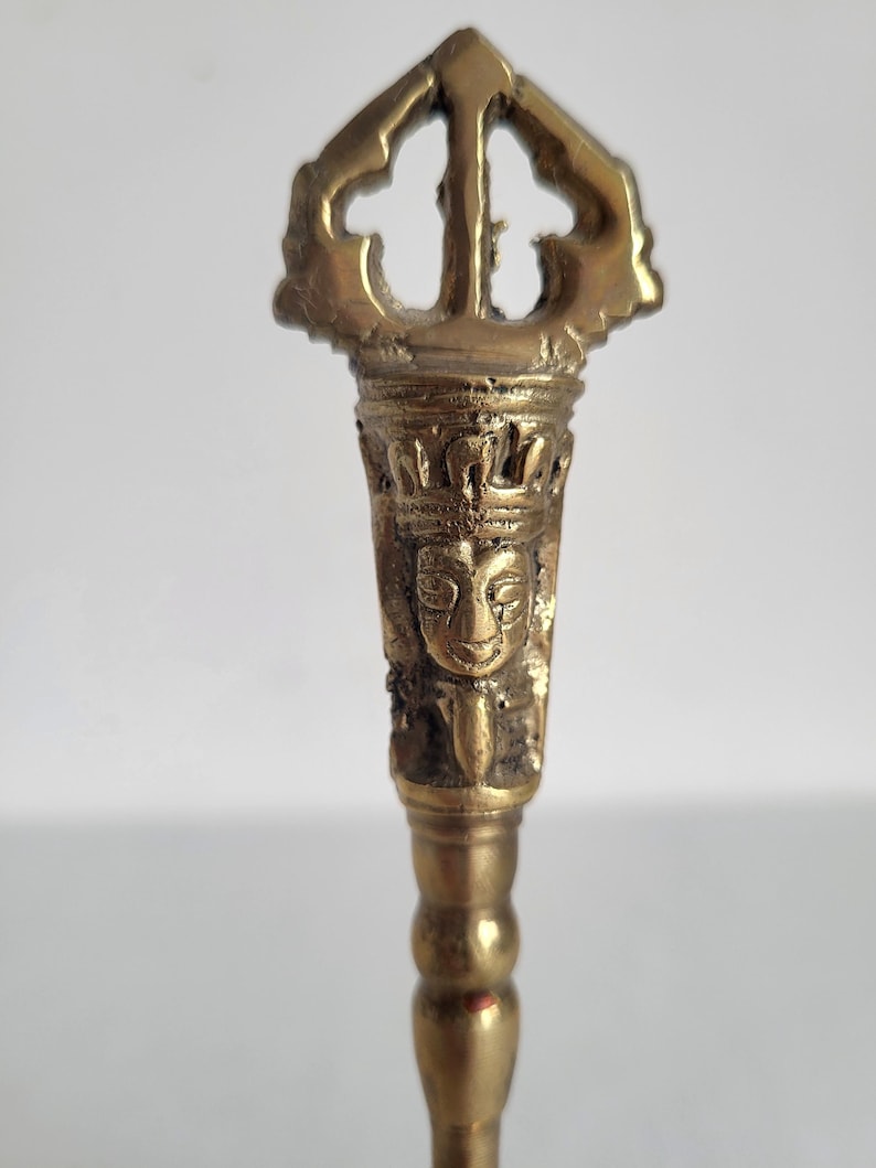 May include: A detailed, antique brass vajra, a ritual object in Buddhism and Hinduism. The vajra features intricate carvings, including a face and ornate crown. The handle has a series of rounded sections, and the top has a three-pronged design.