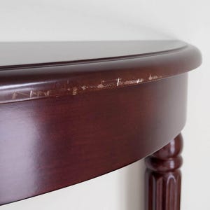 Op de afbeelding: Een gedeelte van een donker kersenkleurig gebeitste houten tafel met een afgeronde rand en een enkele, gecanneleerde poot. Het tafelblad heeft een glanzende afwerking en er zijn zichtbare krassen langs de rand. De tafel lijkt een halfrond ontwerp te hebben.