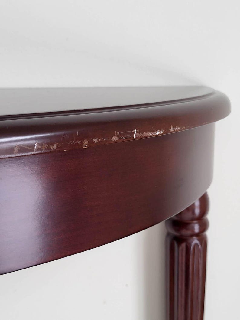 Op de afbeelding: Een gedeelte van een donker kersenkleurig gebeitste houten tafel met een afgeronde rand en een enkele, gecanneleerde poot. Het tafelblad heeft een glanzende afwerking en er zijn zichtbare krassen langs de rand. De tafel lijkt een halfrond ontwerp te hebben.