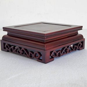 May include: A dark red wooden display stand, square in shape, with a carved openwork pattern around the base. The top surface is flat and slightly recessed. Designed to elevate and display decorative items.