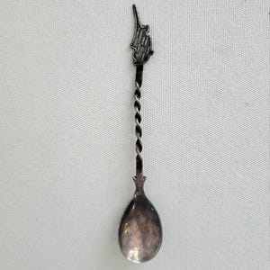 May include: A silver-toned spoon with a twisted handle and a teardrop-shaped bowl. The top of the spoon features a small, detailed emblem. The spoon is likely a collectible or souvenir item.