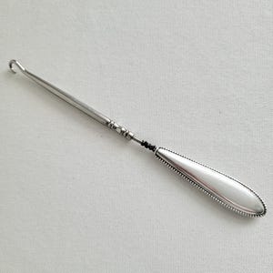 May include: A silver-colored crochet hook with a curved hook end and a long, slender shaft. The handle is oval-shaped with a beaded border. The hook is designed for crafting and textile work.