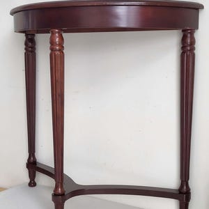 Op de afbeelding: Een mahoniehouten console met halve maan. De tafel heeft een gebogen bovenkant die wordt ondersteund door drie gefreesde poten en een gebogen basis. De afmetingen van de tafel zijn ongeveer 76 cm breed, 38 cm diep en 76 cm hoog.