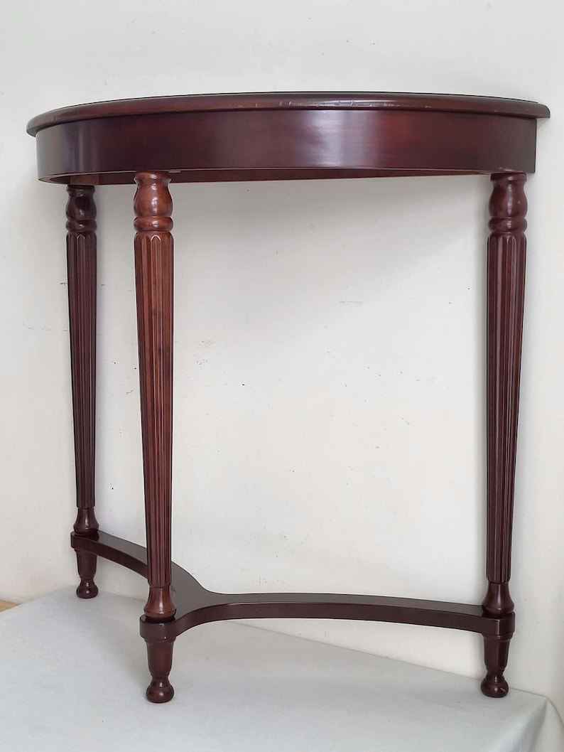 Op de afbeelding: Een mahoniehouten console met halve maan. De tafel heeft een gebogen bovenkant die wordt ondersteund door drie gefreesde poten en een gebogen basis. De afmetingen van de tafel zijn ongeveer 76 cm breed, 38 cm diep en 76 cm hoog.