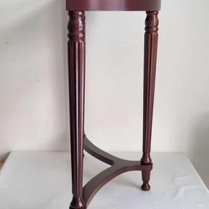 Op de afbeelding: Een donker kersenkleurige, halvemaanvormige accenttafel. De tafel heeft een afgerond blad, ondersteund door drie gefreesde poten die zijn verbonden door een gebogen ligger. De poten hebben decoratieve details.