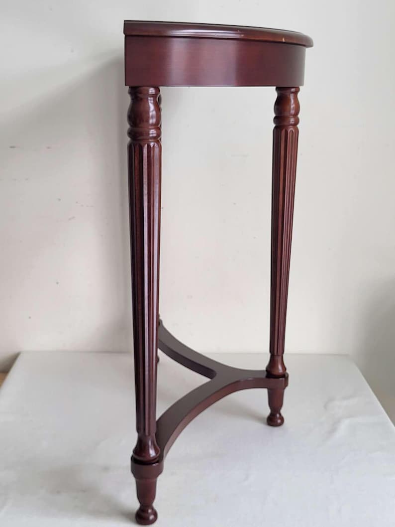 Op de afbeelding: Een donker kersenkleurige, halvemaanvormige accenttafel. De tafel heeft een afgerond blad, ondersteund door drie gefreesde poten die zijn verbonden door een gebogen ligger. De poten hebben decoratieve details.