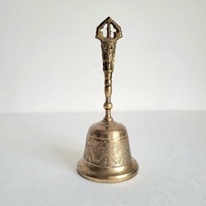 May include: A vintage brass bell with an ornate handle and detailed carvings. The bell has a flared base and a long, slender handle with a decorative top. The bell is likely used for ceremonial or decorative purposes.