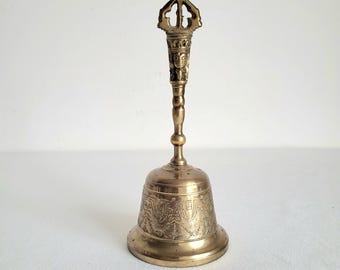 May include: A vintage brass bell with an ornate handle and detailed carvings. The bell has a flared base and a long, slender handle with a decorative top. The bell is likely used for ceremonial or decorative purposes.