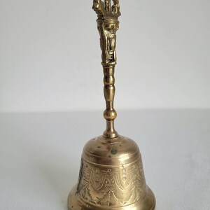 May include: A golden-colored bell with an ornate handle and a detailed, patterned design on the bell's body. The handle features intricate carvings, and the bell has a classic shape. The bell is made of metal.