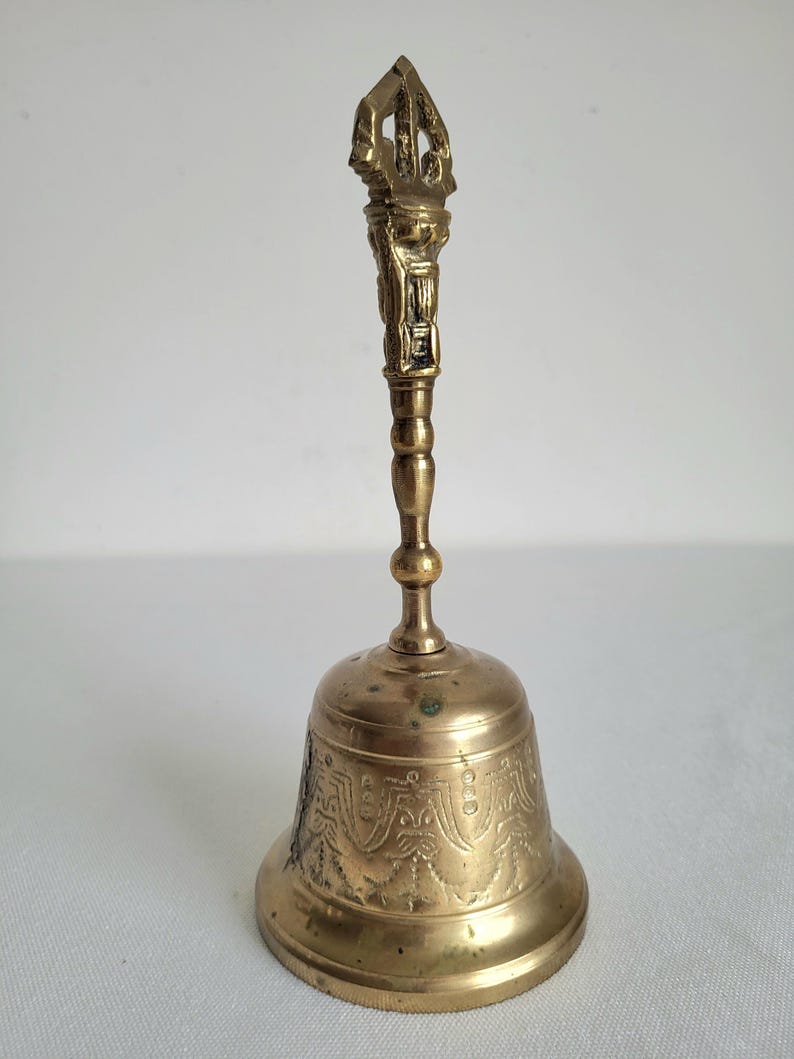 May include: A golden-colored bell with an ornate handle and a detailed, patterned design on the bell's body. The handle features intricate carvings, and the bell has a classic shape. The bell is made of metal.